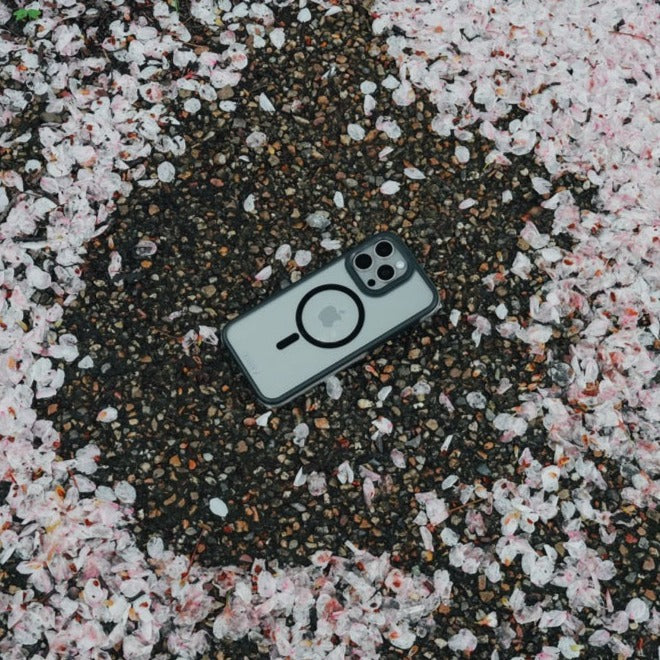 An iPhone 15 Air Plus with three cameras lies among scattered cherry blossom petals on a gravel surface, featuring Military Spec drop protection.