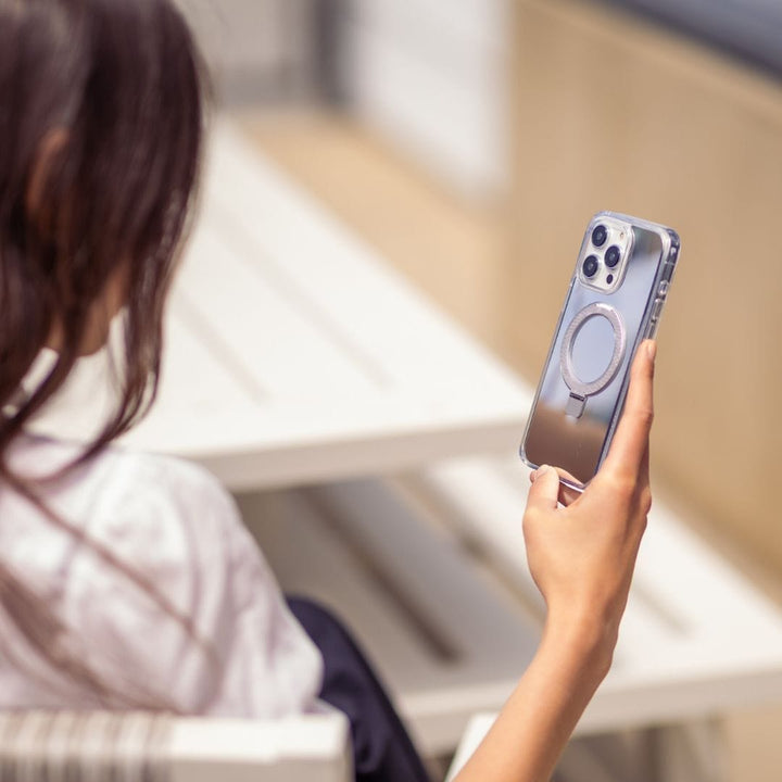 A person with long dark hair, wearing a white shirt, is seated on a white bench outdoors and holding an iPhone 16 Pro Max equipped with MagSafe. The phone is encased in the Raptic Air Stand Mirror Case with a distinct circular design on the back. The blurred background draws attention to the person's hand and the TrueReflect mirror finish of the Raptic case.