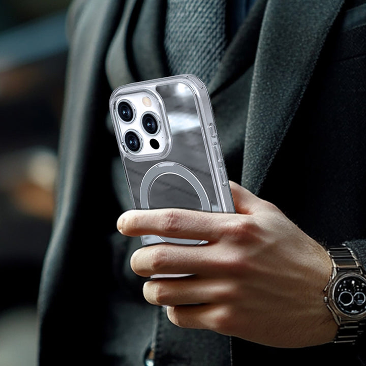 An individual in a grey suit holds a Raptic Air Stand Mirror Msafe iPhone 16 smartphone with a reflective case. The phone features three rear cameras arranged in a triangle. The person is also wearing a wristwatch with a metal strap, and the background is blurred.