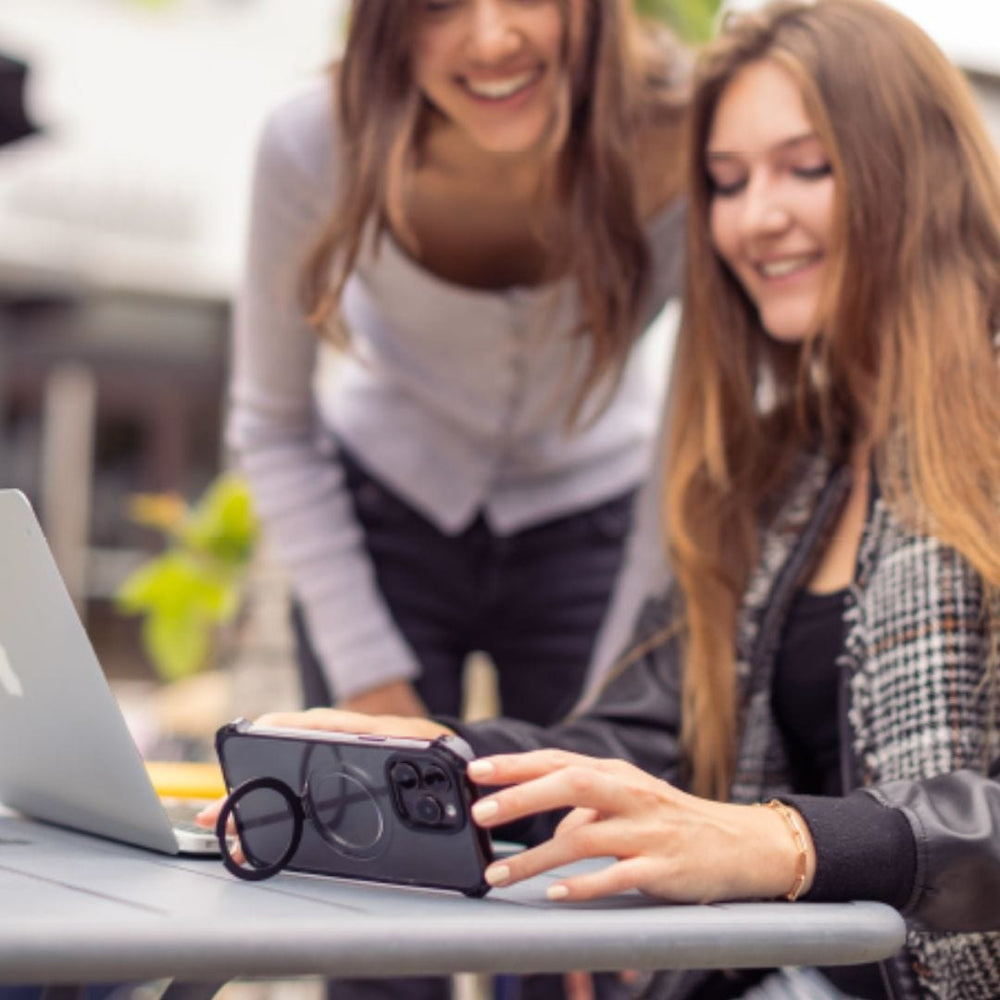 Two women are interacting outdoors. One sits at a table, smiling and holding an iPhone 16 encased in a stylish Raptic Dual Impact Kickstand Case, while the other leans in, also smiling. A silver laptop sits open on the table in front of them. The scene appears casual and friendly, possibly involving shared technology or social media browsing.