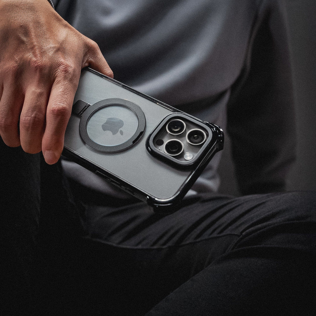 A person holds an iPhone 16 Pro Max encased in a Raptic Dual Impact Kickstand Case, which is MagSafe-compatible and features a circular magnetic ring on the back. The phone showcases three camera lenses arranged in a triangular layout. The person is seated, wearing black pants and a gray shirt, with their hand prominently visible in the image.