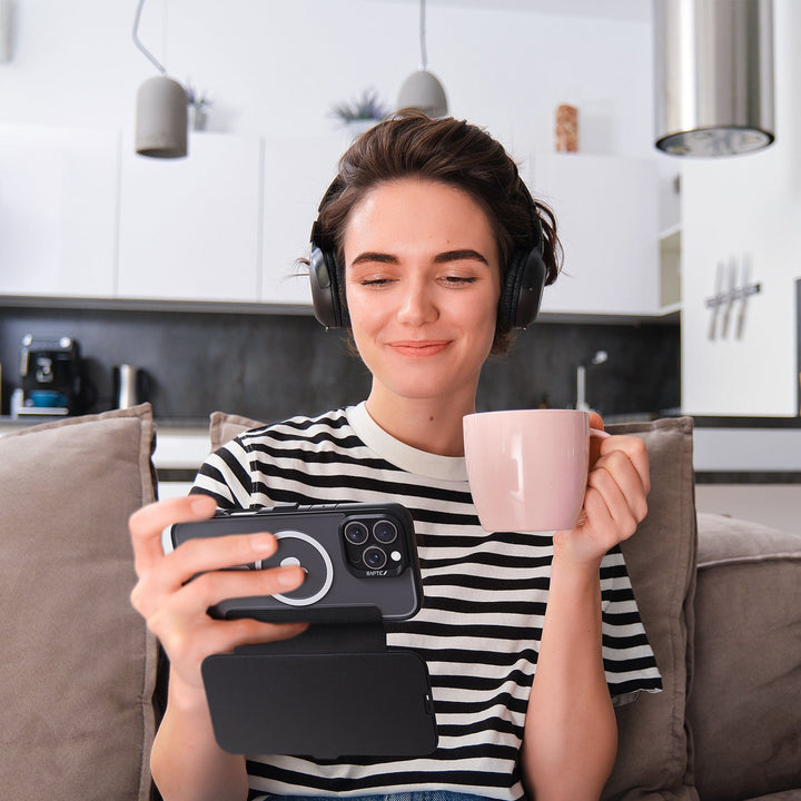 A person with short hair, wearing headphones and a black-and-white striped shirt, sits on a beige couch in a modern kitchen. They are holding the Raptic MagSafe Urban Folio Case with Card Slot for iPhone 16 in one hand and a pink mug in the other, smiling as they look at their phone. Appliances and shelves are visible in the background.