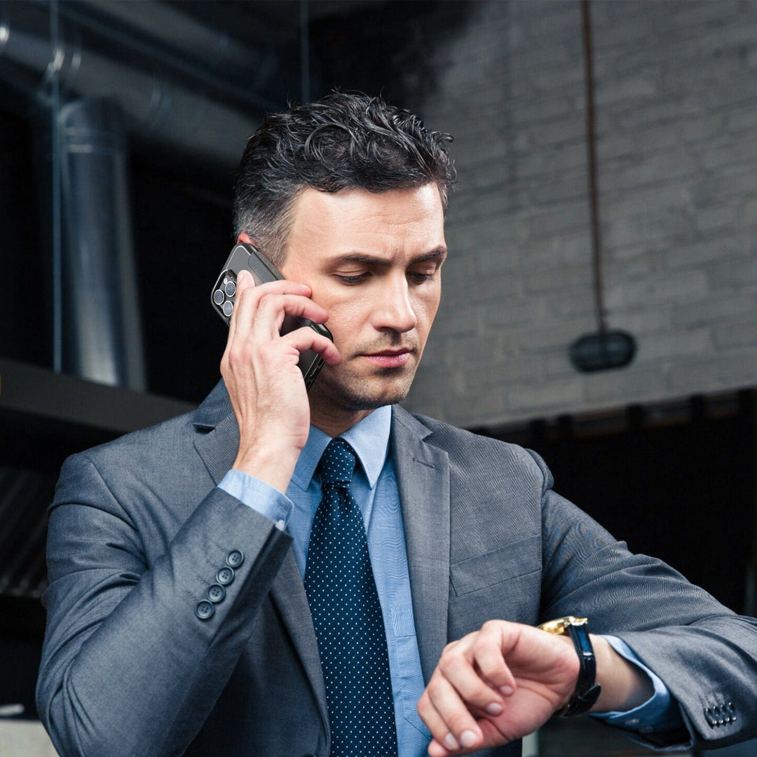 A man in a gray suit, blue shirt, and dark patterned tie is talking on his smartphone with a Raptic MagSafe Urban Folio Case with Card Slot for iPhone 16 while checking his wristwatch. He has short, dark, slightly wavy hair. The background features a brick wall and industrial decor, including exposed pipes and hanging lights.