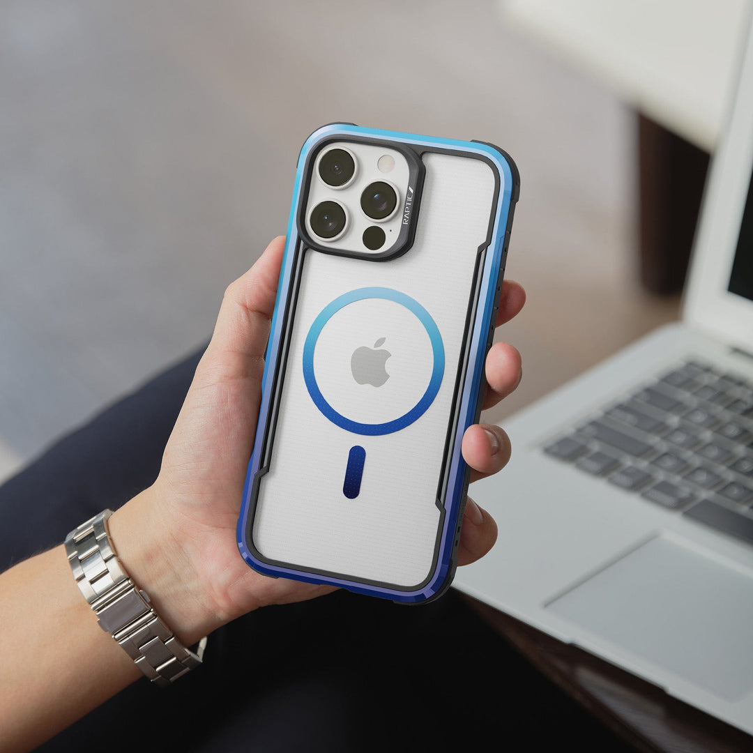 A person is holding an iPhone with a Raptic Shield 2.0 MagSafe for iPhone 16 Pro Max case. The Raptic case is white with a blue outline around the circular MagSafe ring, and the top part of the case accommodates the phone's camera setup. The person is wearing a silver watch and sitting in front of a partially visible laptop.