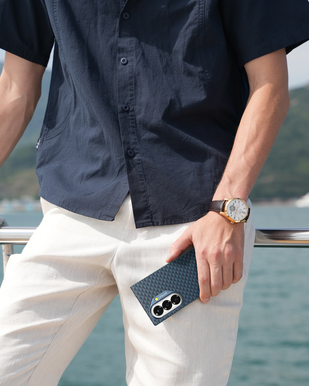 A person in a navy short-sleeve shirt and beige pants holds a RAPTIC Samsung Galaxy Fold 7 Skin Case - Profound Navy by the railing, wearing a watch with a white face and brown strap. Water and blurred hills appear in the background.
