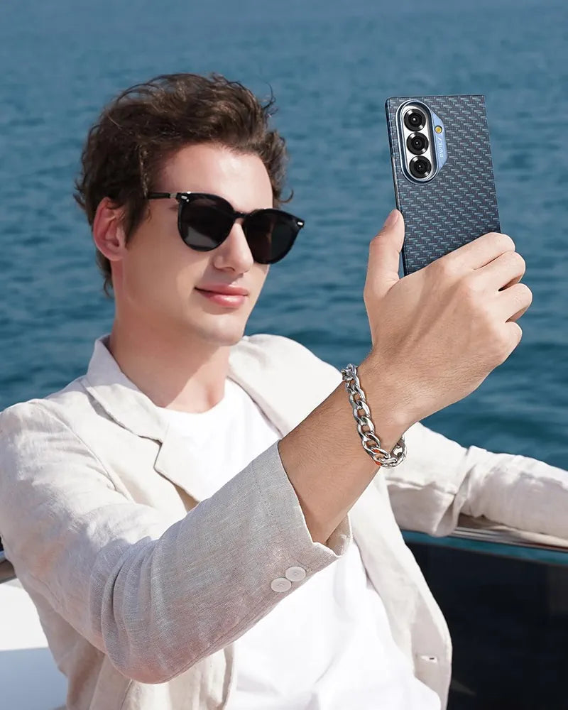 A young man in sunglasses and a light blazer smiles as he takes a selfie on a boat using his phone, protected by the RAPTIC Samsung Galaxy Fold 7 Skin Case in Profound Navy. The blue ocean sparkles behind him, and he wears a chunky silver chain bracelet.