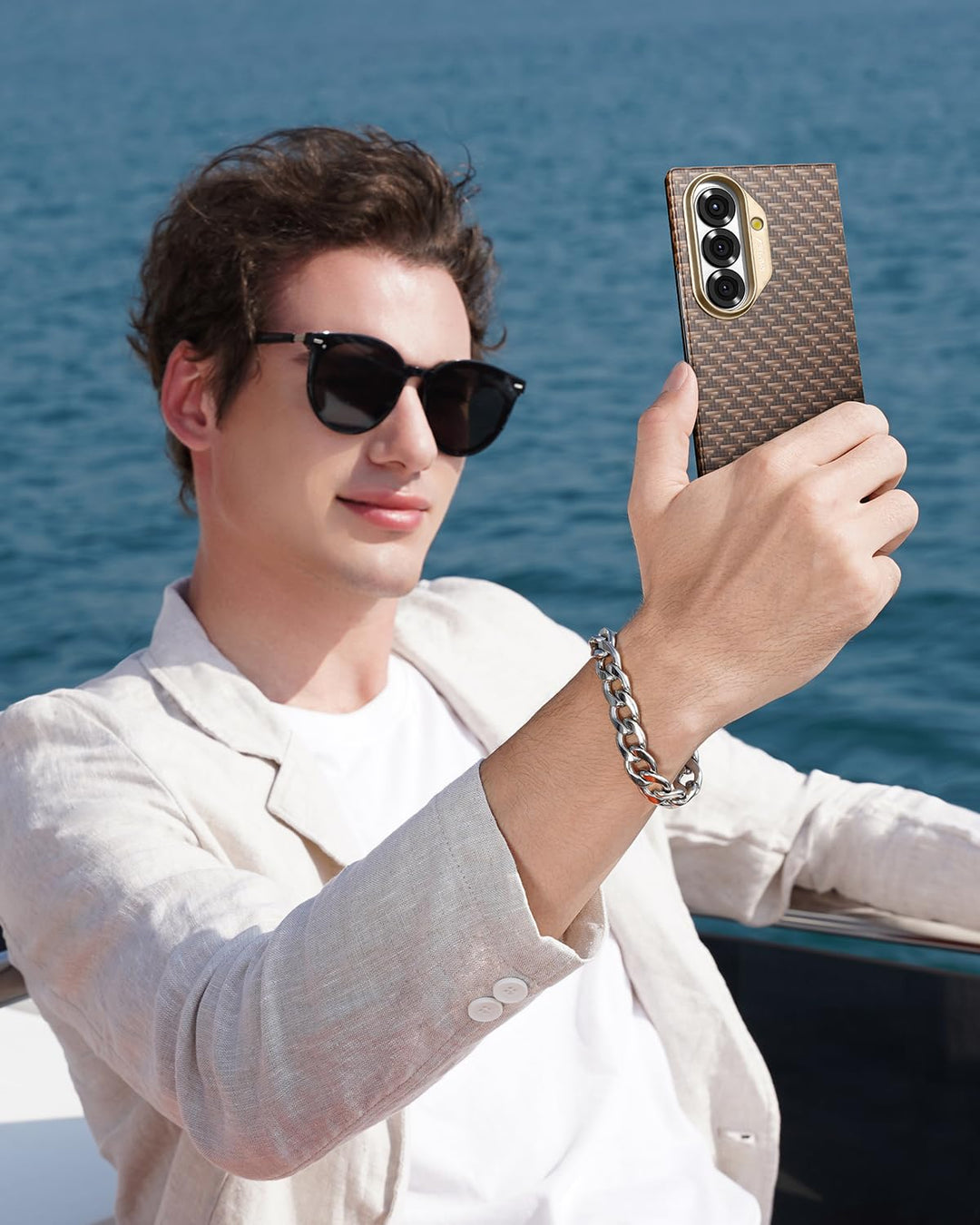 A young man with short brown hair, in black sunglasses and a beige blazer, takes a selfie on a boat with his Samsung Galaxy Fold 7 in the RAPTIC Humble Desert Skin Case, the blue ocean stretching out behind him.