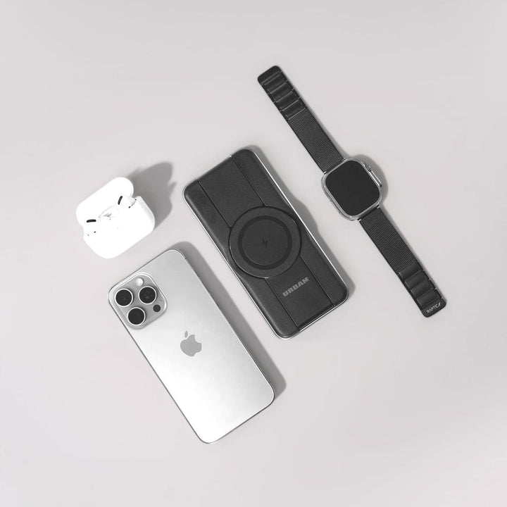 A silver iPhone with three cameras is placed next to a pair of white wireless earbuds in an open charging case. Beside the Urban PowerBuddy 5in1 Wireless MagSafe Powerbank 2 and a black Apple Watch with a black band, this travel-friendly setup boasts universal compatibility and is set against a plain, light-colored background.