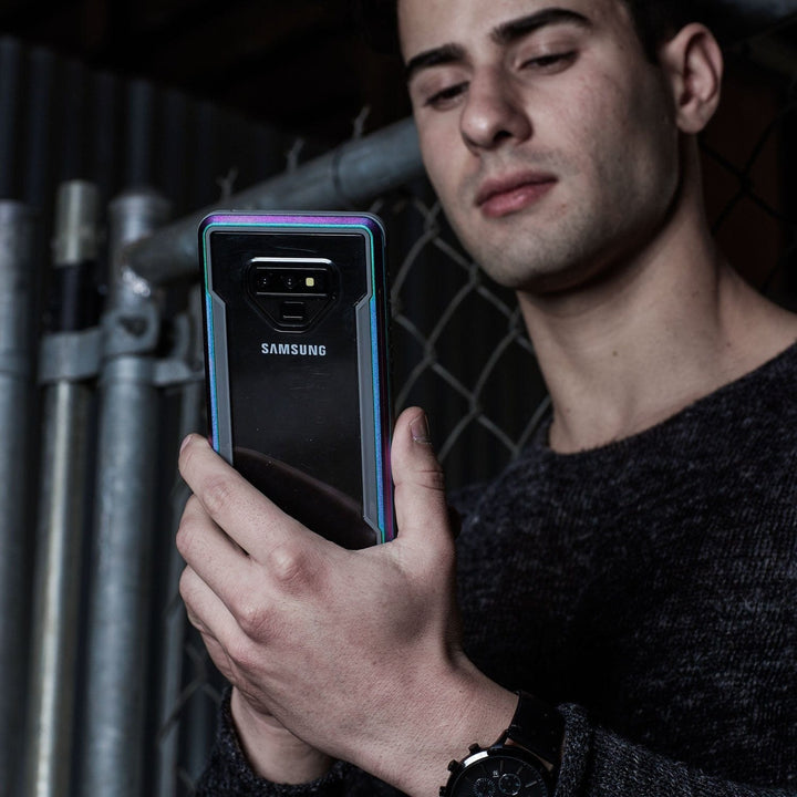 A person in a dark gray sweater holds a Samsung Galaxy Note 9, encased in an X-Doria Defense Shield case by Raptic, in front of a chain-link fence.